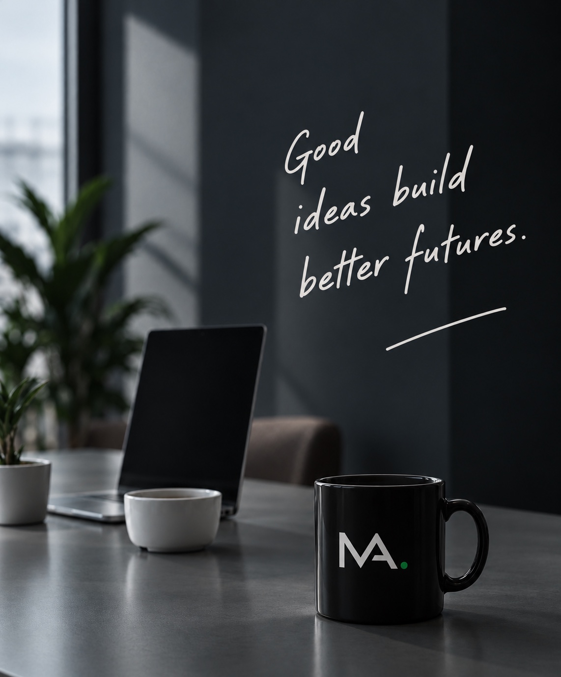 A desk with a laptop, plant and MA mug, beside the words Good ideas build better futures